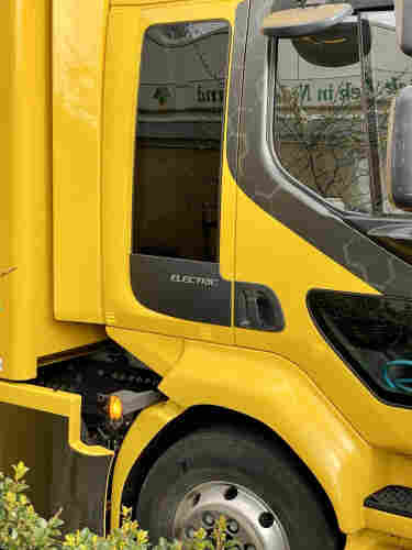 A close-up view of a yellow electric truck cab, highlighting the 'ELECTRIC' label and the side mirror reflecting building windows.