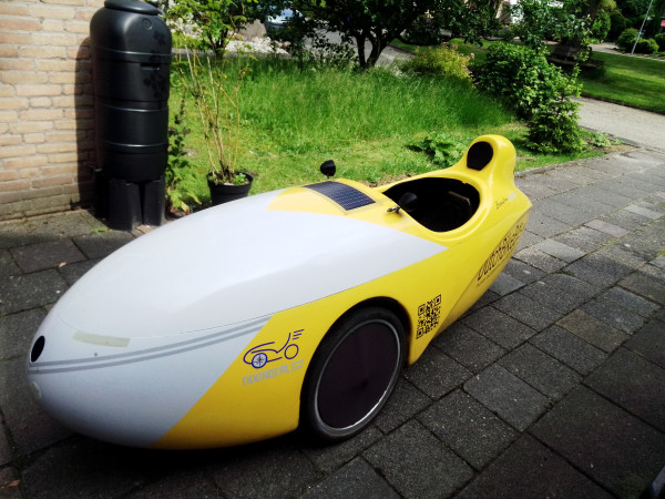 A picture of my Mango velomobile relaxing on our driveway after returning from this morning's ride. There's a water butt and an unkempt garden behind (we do "no mow May," as well as other months ;-)