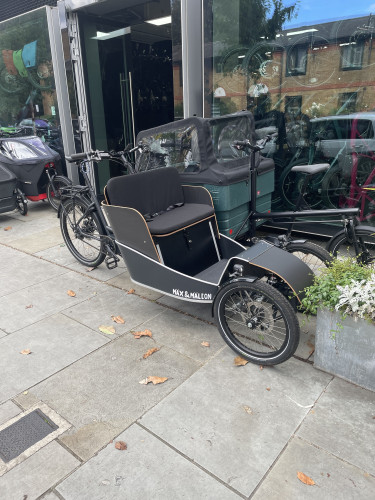 An electric cargo bike with one back wheel and two articulated front wheels is on a pavement outside a shop. It’s got an upright passenger seat like a rickshaw just in front of the handlebars.