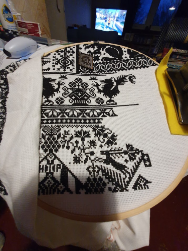 Black and white cross stitch in a wooden hoop. The pattern is busy, lots of designs going on - a partial dragon, a hare, what could be flowers. My messy desk filled with pills, controllers, polish can be seen. The tv is on in the background with Formula One testing on it. 