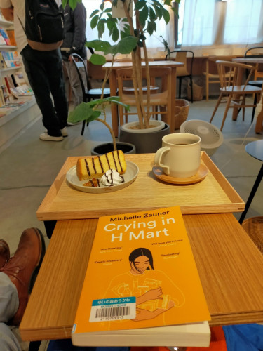 The photo is of inside the cafe. The orange paperback library book has an illustration of a young Asian woman with black her hands full of white outlined foodstuffs. Behind it is a light brown wooden tray. On the left of the tray is a piece of chiffon cake perpendicular on the plate with a small pile of orange rinds and on the right a dollop of whipped cream, all 3 of which of stripes of chocolate on them. To the right is a grey mug of black coffee on a small round wooden coaster. In the distance you can see a green plant, wooden tables and chairs and 2 headless men standing, one with a backpack