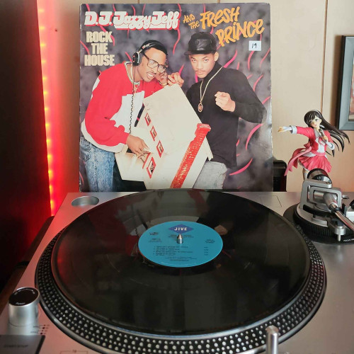 A vinyl record sits on a turntable. Behind the turntable, a vinyl album outer sleeve is displayed. The front cover shows Jazzy Jeff holding a toy house while wearing headphones. Will Smith is posing and pointing at the camera. 