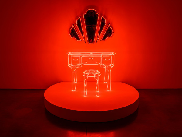 Neon sculpture of a vanity (mirror, desk, and stool) against a red wall