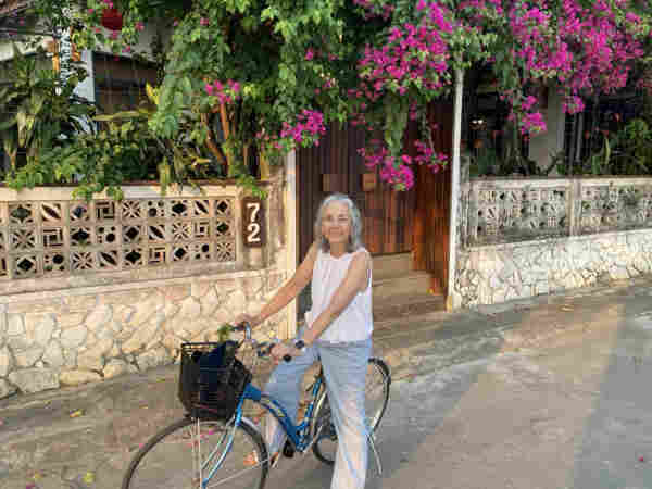 A grey haired woman sits astride a bicycle with a basket in front of a villa with abundant bougainvillea overhanging, spilling over the street. 