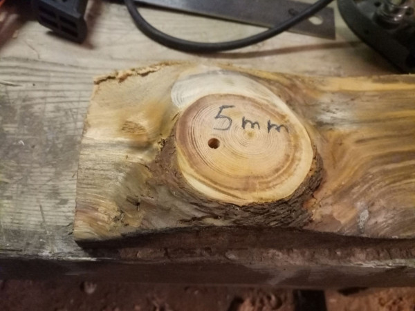 Top of a piece of firewood with a massive knot in it and a flat sawn bottom to rest against a workpiece. A hole is drilled through the knot (not in the center, because the knot exits at an angle) and pencil marked "5 mm".