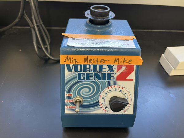 Machine for swirling test tubes to mix them (Vortex Genie 2: a blue box with a toggle switch and speed dial on the front and a cupped moving unit on top) with an orange tape label on which “Mix Master Mike” is written in black sharpie. Sitting on a black epoxy resin lab bench with lab detritus scattered nearby