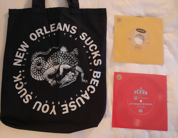 A black tote bag that says "New Orleans Sucks Because You Suck" from Sisters in Christ record shop. Next to bag are two 45 records, a yellow one by the Meters. And a red one by Eyehategod (pronounced Eye Hate God)