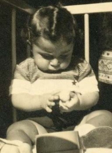A vintage sepia-toned photo showing a baby seated, focusing intently on their hands as they play with a small object. The baby wears a striped outfit and has neatly combed hair with a visible part. 