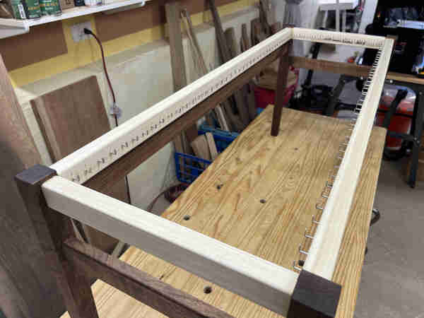 The top of a partially completed sitting bench. It does not have a seat yet. The rails are light colored and the legs are dark. There is a single row of nails running around the entire inside of the rails where the weaving for the seat will attach.