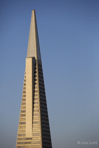 A Simple Colour Photo In Portrait Format. The Image Shows The Upper Section (About 21 Floors) Of A Sharply Pointed High-Rise Building. The Building Is Isolated Against A Clear And Cloudless Sky. The Building Has Two Sides Visible With Two Further Sides Behind And Out Of Shot. On The Camera Side Is A 'Wing', A Wedge Shaped Outcrop That Houses The Lift Shaft That Services The Upper Floors. On The Top Of The Building Is A Point, Perhaps A Lightening Conductor. 
The Building Is The Iconic Transamerica Pyramid, Once The Tallest Building In San Francisco.

San Francisco, California. 2013