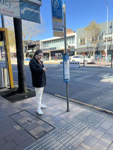 A person standing at a bus stop, looking at their phone. The bus stop sign indicates routes to Neutral Bay Junction, Mosman, Northern Beaches, and Taronga Zoo. There are shops and buildings in the background.