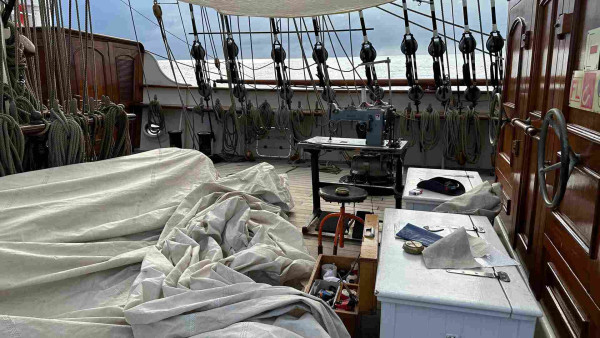 Sewing machine and sail spread across the foredeck of a ship with pin rails and a mast in the background. 