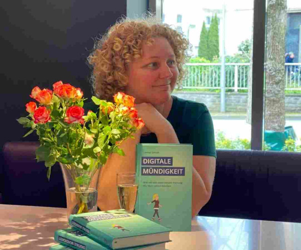 Leena Simon sitzt stolz bei der Feier ihres Bucherscheinens hinter ihrem Buch "Digitale Mündigkeit" einer Vase mit hellroten Rosen und einem Sekt-Glas.