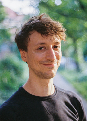 An analog portrait photo of a young man, looking straight into the lens, standing slightly sideways to the camera. He smiles, has dark brown hair and a dark blue shirt.