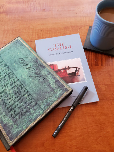 Poetry collection The Sun-fish by Eiléan Ní Chuilleanáin (The Gallery Press), with its bright blue cover, sits on the dining room table with a notebook with an ornate green cover, a black pen and coffee in a blue cup