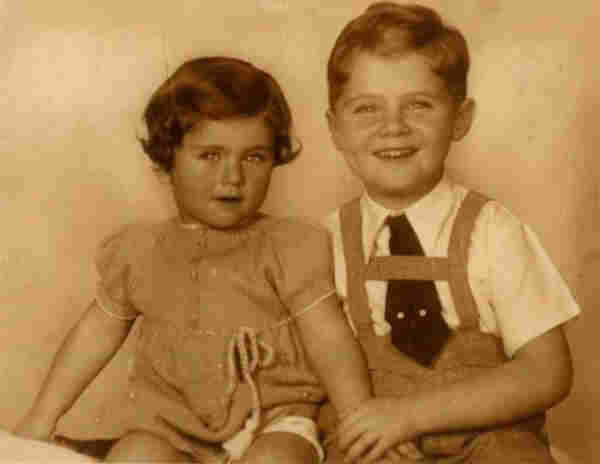 Photo of a girl and a boy. They are sitting next to each other. The boy is holding the girl's hand. Both are smiling. The girl is wearing a dress, the boy pants with suspenders and a tie.