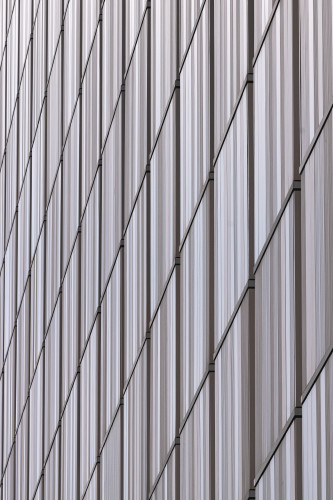 Color photograph of one of the exterior facades of the University of Iowa School of Music (Voxman Building). The facade's surface is paneled and made up of vertical and diagonal elements. The vertical lines of each panels are made up of slender grey, brown-grey, brown, and black panels   that change colors each time they cross a diagonal element. Each panel is framed by three-dimensional, vertical support columns as well as the three-dimensional diagonal elements. Both the vertical and diagonal framing are the dark grey color. The photo was take at an oblique angle. In reality, the diagonal elements are actually horizontal.

