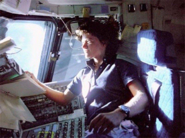 Sally Ride in the Challenger cockpit during STS-7. She is in a seat but also appears to be floating. Sunlight reflected off the Earth is streaming in through the windows and illuminating her. There is a bank of switches and dials in front of her, as well as a manual or book of some sort. Ride is looking out the window, squinting slightly because of the bright light.