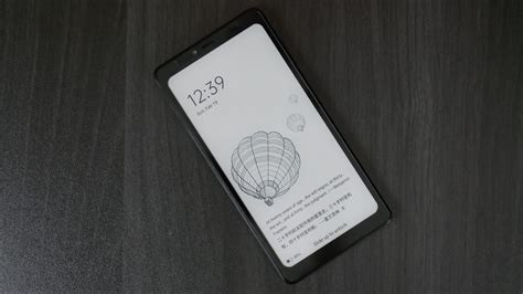 A picture of an e-ink display smart phone on a dark-grey stained wooden surface. The phone has the time, an image of a hot air balloon, and some small text displayed on the screen.