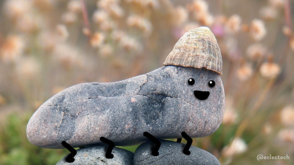 A photo of a pebble, grey with speckles of pink running through it. It is short and wide, and sits on top of two rounder grey pebbles. The backdrop is blurred seaside thrift flowers. On the right hand side of the pebble there is a limpet type shell like  a beanie hat. I have drawn a simple happy face below the shell / hat and multiple short legs.