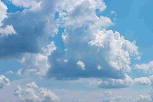 Photograph of a vivid blue sky filled with towering banks of cumulus clouds. The clouds are highlighted on their right side by the sun and are in shadow on the left side. In the lower left frame there is a tiny black helicopter that is dwarfed by the clouds around it.