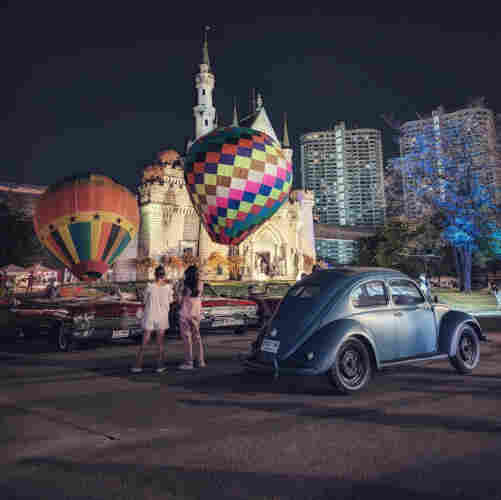 Two girls standing in front of a row of classic cars and admiring the view of colorful hot air balloons. Just behind the hot air balloons is a well lit castle, with doors wide open, welcoming visitors to go to the upper level for views of the city and night market