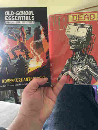 A person is holding two books: one titled "OLD-SCHOOL ESSENTIALS OFFICIAL ADVENTURE SCENARIO" featuring an armored figure in a dynamic pose, and the other titled "VR DEAD" with an illustration of a skeletal figure wearing a VR headset.