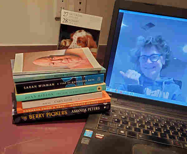 Silent book club member Vicki (me) is visible on her computer screen, getting ready for a silent book club zoom meeting. Next to the computer is a dog calendar and some books, including works by Kirby, Matthew Tierney, Sarah Winman, Joan Didion, Anik See, Bradley Peters and Amanda Peters.