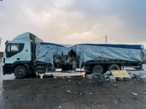 A damaged truck with a tarp covering its cargo area, parked on a paved surface under a cloudy sky. Debris is scattered around the truck.