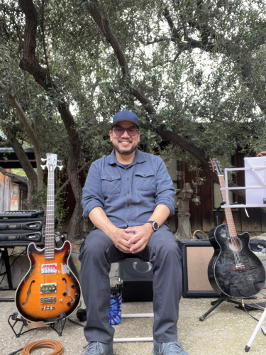 A person in casual outdoor attire sitting on a stool between two guitars under a tree. Musical equipment is visible in the background.