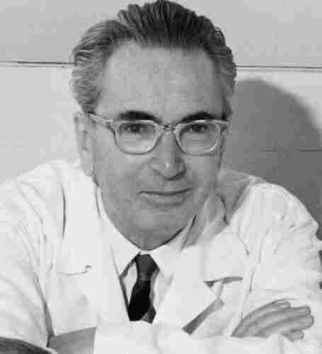 A mature man in a white shirt, dark tie and an outfit that resembles a doctor's kit. He is wearing thick-rimmed glasses. His eyebrows are wide and his hair is pulled back. His arms are folded.