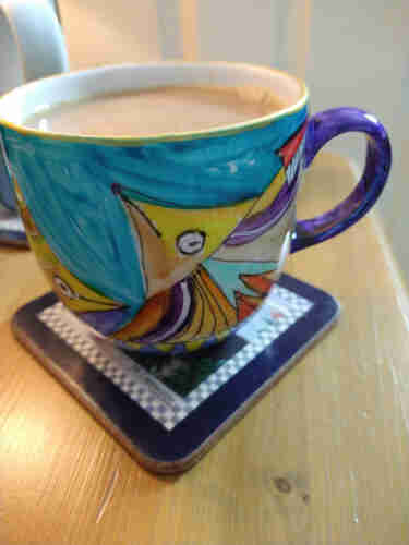 Colourful mug of tea on a bedside cabinet there is what looks like a fish on a blue background it has a yellow triangular shaped head and multicoloured body 