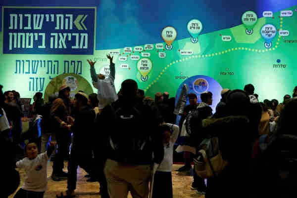 A crowded event with people gathered in front of a large graphic display with Hebrew text saying “settlements bring security”