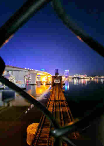 From a pedestrian overpass encircled by thin black chain link wire caging, the view thru one diamond shaped opening directly above a train track crossing a wide river. The railway drawbridge is down in anticipation of an approaching train. Beneath a dark, deep blue night sky, a nearby vehicle bridge is colorfully illuminated, with the far shoreline lined with colorfully lit buildings, all casting reflections upon the calm water below.