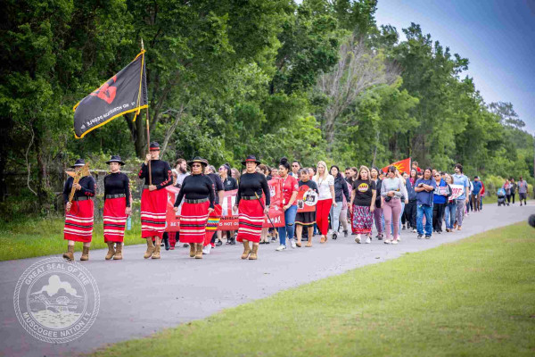 The Muscogee Nation MMIP Honor Walk headed by five Mvskoke women in red ribbon skirts with black shirts, followed by a crowd of Native people marching.
