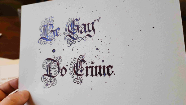 'Be Gay Do Crime' calligraphed in dark blue sparkly ink with lots of ink splatters and very ornate majuscule letters