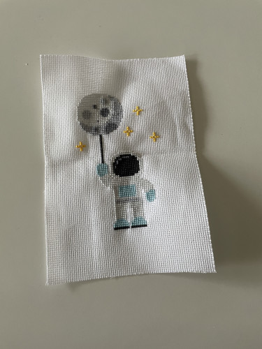 Cross stitch on a white cloth of an astronaut in a spacesuit, holding a moon on a string like a balloon, surrounded by a couple of stars.