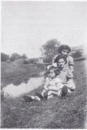 Three children sitting by a pond in a grassy area, with houses visible in the background.
