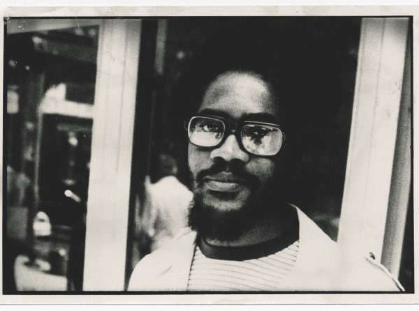 Black & white photo of Walter Rodney, a dark skinned Black man with a beard & moustache & middle length natural hair. He's wearing glasses and a light colored jacket over a t-shirt. His expression is flat.