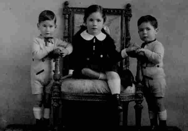 Picture of three kids. In the middle, in an arm-chair sits a gul in a dress. On both sides of the chair her twin brothers are standing.