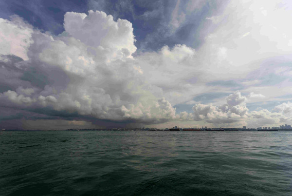 view of the Singapore Straiti from a moving boat, with a dull green sea in the foreground, a line of buildings and port structures on the horizon and a massive burst of clouds in the sky above.