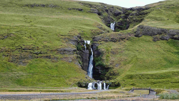 A photo of a waterfall in summer, with white water dropping from the top of a grassy green bank. There are three actual drops - one is visible at the top of the shot, one in the centre and the final one almost at ground level. In the foreground on the right is a small road bridge across the water flowing away from the falls, a car parking area is next to it and one vehicle is parked near the waterfall. A few people can be seen climbing a gravel path and steps along one side of the drops, giving the scene scale.