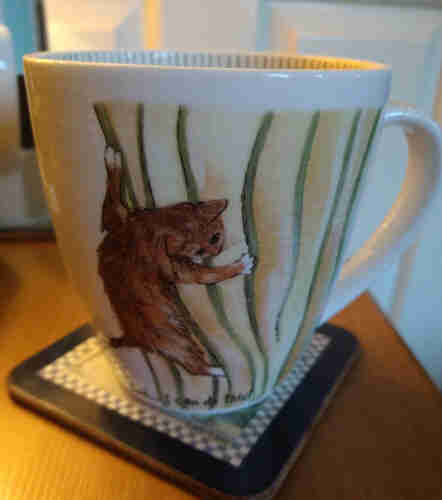 Large mug of tea on a bedside cabinet, it has an image of a ginger cat climbing up yellow and green curtains 
