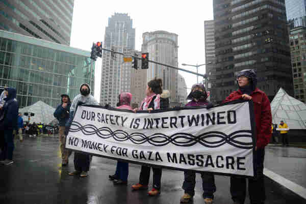 Activists on a rainy street in a downtown area, holding a banner that says “Our Safety is Intertwined / No More Money for Gaza Massacre”. Running horizontally across the middle is an ornamental “intertwined” pattern of two threads looping around one another, almost like links in a paper chain or a double helix.