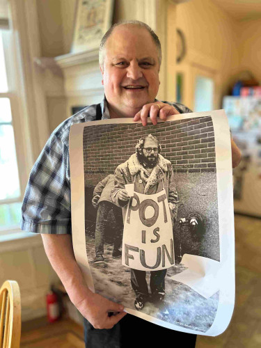 Steve in a blue checked shirt holding an image of Allen Ginsberg holding a poster that says POT IS FUN with snow dusting his hair.