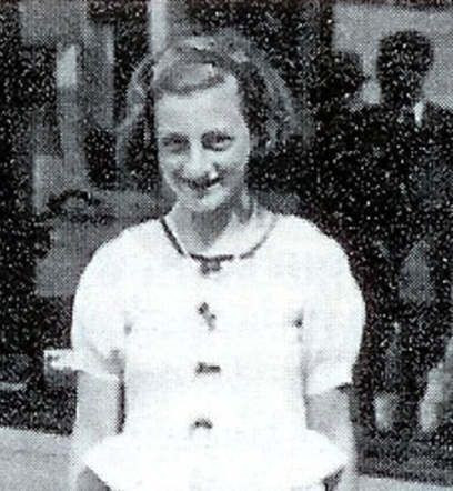 Old black and white photo of a smiling young girl in a buttoned dress, standing outdoors.