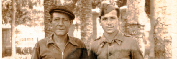 Two men stand together in front of what appear to be the trunks of four palm trees. The man on the left is older, in a worker’s flat cap and unzipped kacki  shirt.  The man on the right appears to be his son, he has a pointed military cap on, it is cocked to the right. He had a buttoned uniform. They have nearly identical expressions. 