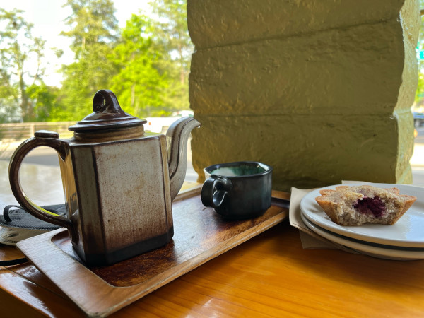 Tall rectangular crème & brown glazed ceramic tea pot with small handled tea cup in black and teal. A half eaten raspberry tart is on a plate. The ensemble is on an outside bar top and it’s golden hour 