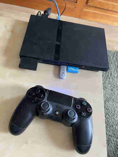 Una consola PlayStation 2 Slim enchufada con un cable de red, una tarjeta de memoria, un pendrive USB azul y un adaptador Bluetooth. Al lado hay un mando Dualshock de PlayStation 4. 