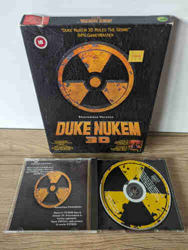 Photo of the game box and CDROM for the game "Duke Nukem 3D" from 1996, shareware version. The box art is some dark metal grey with a large yellow and dark icon for "nuclear warning" also known as atomic symbol in the center and the title in bold yellow letters below. A price sticker says 5,99 GBP Virgin (Store). 
